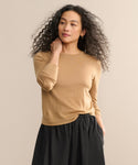 A woman with curly hair models the Mercer Crewneck, styled with a black skirt. She stands against a beige background, gazing to the side with one hand at her neck and the other in her pocket.