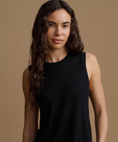 A woman wearing a black sleeveless top, photographed against a solid warm beige studio background.