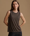 A woman modeling a sleeveless crew-neck tank top in a dark taupe-brown color, paired with a dark pleated skirt, against a solid warm beige studio background.