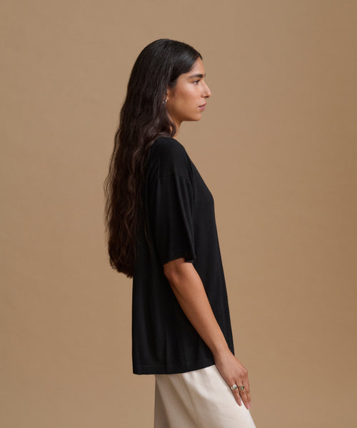 A woman is shown in profile wearing an oversized black short-sleeve top and cream-colored pants, standing against a solid warm beige studio background.