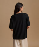 A woman viewed from the back wearing an oversized black short-sleeve t-shirt and cream-colored pants, set against a solid warm beige studio background.