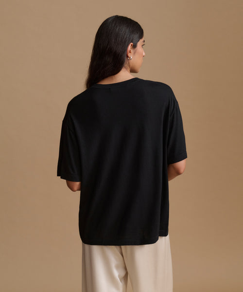 A woman viewed from the back wearing an oversized black short-sleeve t-shirt and cream-colored pants, set against a solid warm beige studio background.
