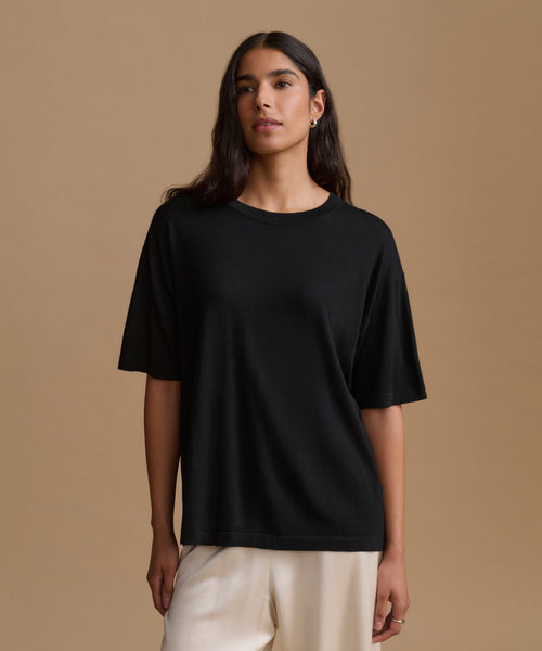 A woman modeling a black short-sleeve crewneck t-shirt with relaxed fit, paired with cream-colored pants, set against a solid warm beige studio background.
