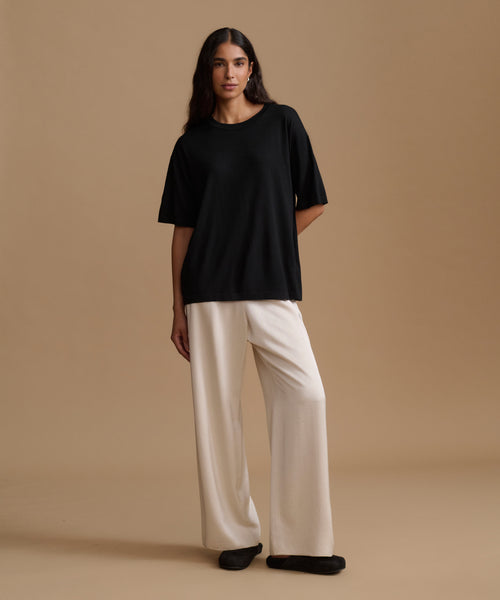 A woman modeling an oversized black short-sleeve t-shirt and cream wide-leg trousers against a solid beige studio backdrop.