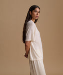 A woman is shown in profile wearing a loose, short-sleeve cream-colored t-shirt and matching cream pants against a solid beige studio background.