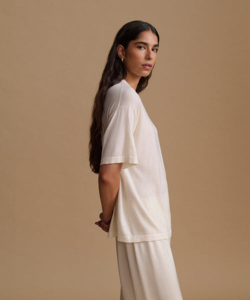 A woman is shown in profile wearing a loose, short-sleeve cream-colored t-shirt and matching cream pants against a solid beige studio background.