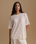 A woman modeling an oversized short-sleeve crewneck t-shirt in a warm off-white/cream color, paired with matching cream pants, set against a solid beige studio background.