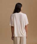 A woman viewed from behind wearing a loose short-sleeve cream-colored t-shirt and matching cream pants against a solid beige studio background.