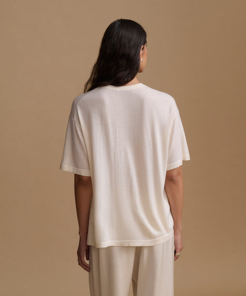 A woman viewed from behind wearing a loose short-sleeve cream-colored t-shirt and matching cream pants against a solid beige studio background.