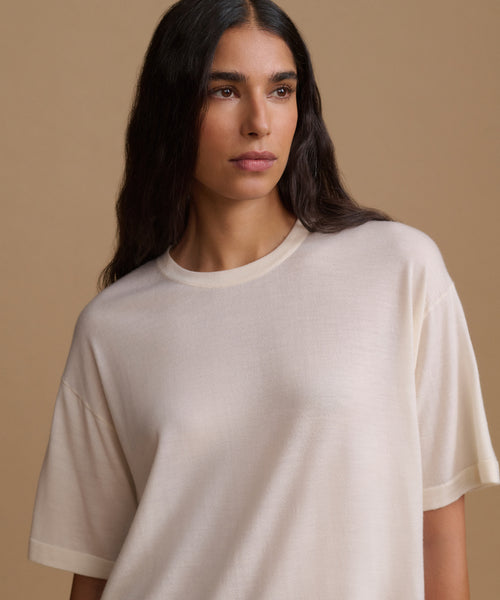 A woman wearing a cream-colored, short-sleeve crewneck t-shirt, photographed against a solid beige background.