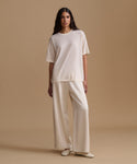 A woman modeling a matching cream-colored set consisting of a short-sleeve crewneck top and wide-leg trousers, paired with cream pointed-toe flats, set against a solid beige studio backdrop.