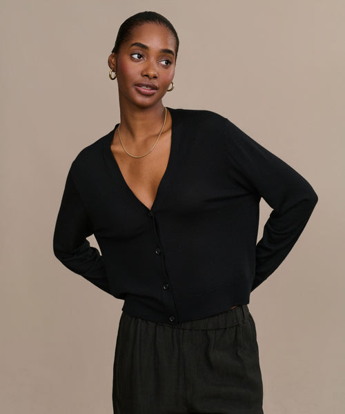 A woman with pulled-back hair wears gold hoop earrings, a thin gold necklace, the Mira Cardigan in black, and dark pants, posing with her arms behind her back against a plain beige background.