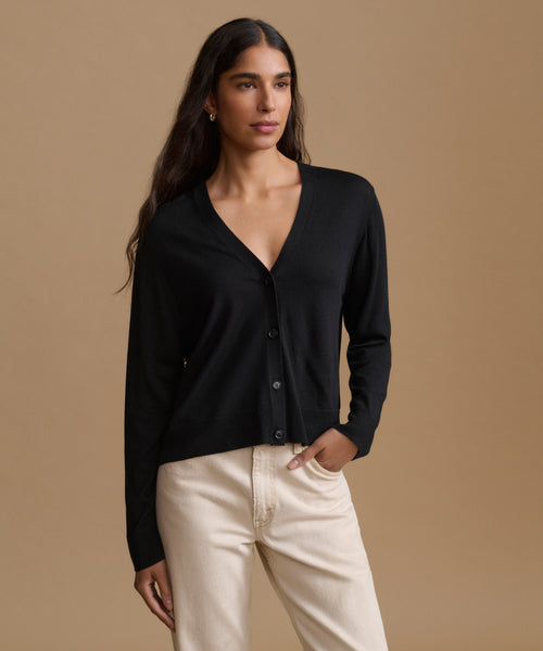 A woman modeling a black long-sleeve button-front cardigan with a V-neck, paired with cream-colored pants, against a solid warm beige studio background.