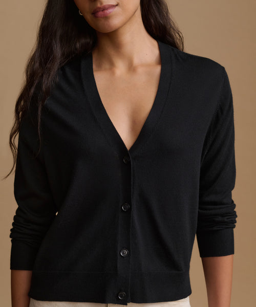 A woman wearing a black long-sleeve V-neck button-up cardigan sweater, shown from the chest up against a solid warm beige background.