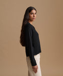 A woman is shown in profile wearing a black long-sleeve cardigan and cream-colored pants, standing against a solid beige studio background.