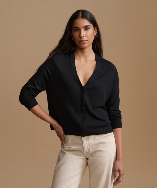 A woman modeling a black long-sleeve button-front cardigan with a deep V-neck, paired with light beige pants, against a solid warm beige studio background.