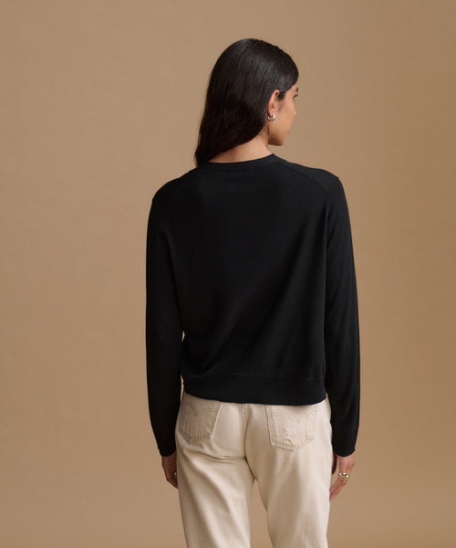 A woman shown from the back wearing a black long-sleeve crewneck sweater and cream-colored pants, set against a solid warm beige studio background.