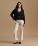 A woman modeling a black long-sleeve button-front cardigan with cream straight-leg pants and black flats, standing against a solid beige studio background.