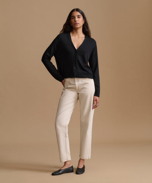 A woman modeling a black long-sleeve button-front cardigan with cream straight-leg pants and black flats, standing against a solid beige studio background.