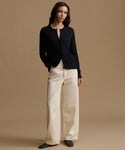 A woman modeling a black long-sleeve button-front cardigan with cream wide-leg trousers, standing against a solid warm beige studio background.