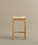 The Montecito Counter Stool features a minimalist design with straight wooden legs and a woven Danish paper cord seat, shown here in a Warm Oak finish against a plain beige background.