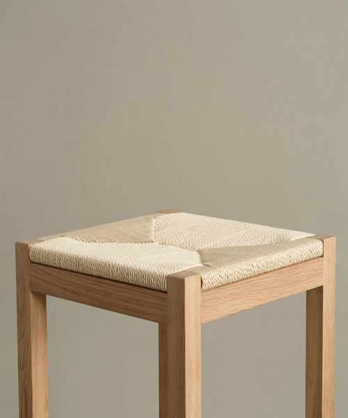 A close-up of the Montecito Counter Stool highlights its woven Danish paper cord seat and minimalist silhouette. Its warm oak finish and clean lines stand out against a plain beige background.