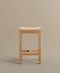 The Montecito Counter Stool features clean lines and a square Danish paper cord seat in warm oak, shown against a plain beige background. Its natural wood finish creates a minimalist, modern look.