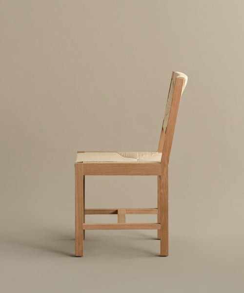 The Montecito Dining Chair, made from warm oak with a handwoven Danish paper cord seat and backrest, is shown side-on against a plain beige background. Its minimalist design features clean, straight lines.