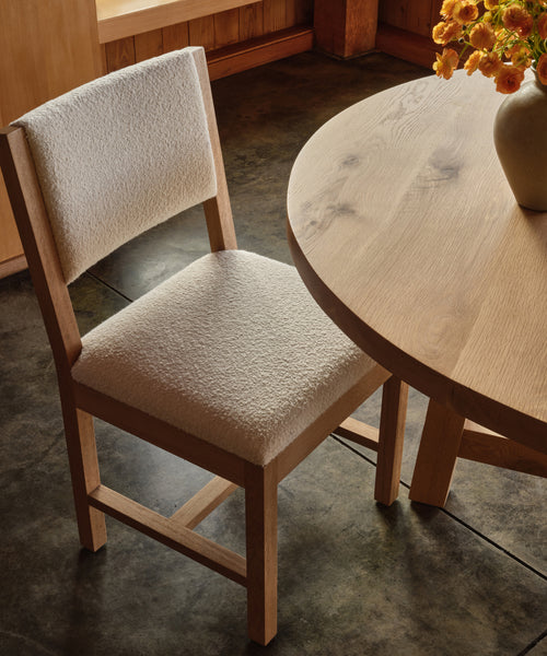 The Montecito Upholstered Dining Chair with ivory boucle upholstery sits beside a round wooden table topped with a ceramic vase of orange flowers, all arranged on a polished concrete floor in a warmly lit room.