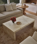A modern living room features the Napa Coffee Table with a vase of red flowers and a plate, set on a light carpet near a cream sofa with light pillows and a round wooden side table.