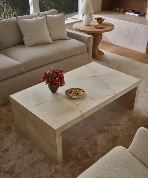 A modern living room features the Napa Coffee Table with a vase of red flowers and a plate, set on a light carpet near a cream sofa with light pillows and a round wooden side table.