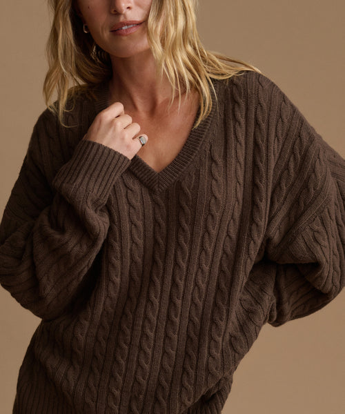 A woman wearing an oversized dark brown cable-knit V-neck sweater against a warm beige background.