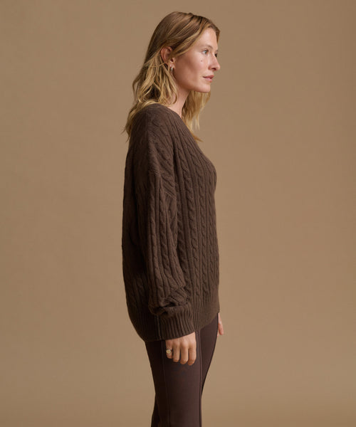 A woman in side profile wearing a dark brown cable-knit sweater and matching dark brown pants, standing against a solid warm beige studio background.