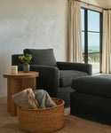 A cozy living room corner features a dark armchair, ottoman, wooden side table with a plant and candle, and a Nesting Basket filled with folded blankets near large windows dressed in beige curtains.