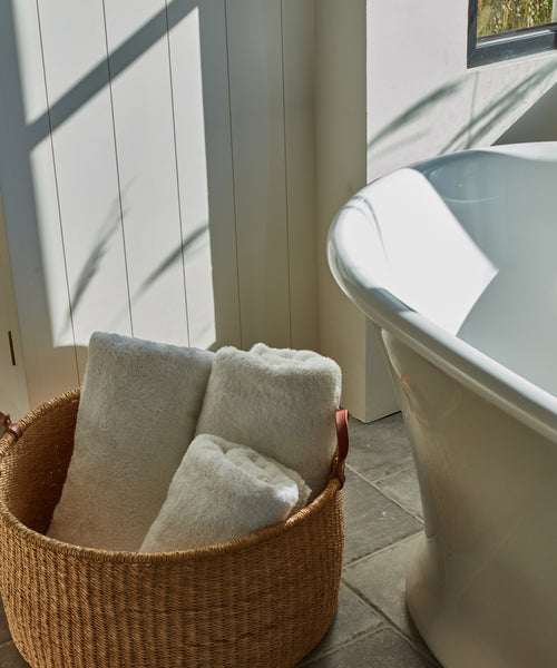 A Nesting Basket made of handwoven elephant grass holds rolled white towels on a tiled floor beside a white freestanding bathtub in a sunlit bathroom.