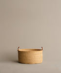 The Nesting Basket, a round handwoven elephant grass basket with brown handles, rests on a plain light beige surface against a matching beige background.