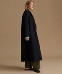 A woman stands in profile wearing a long black coat over olive green wide-leg pants and black flats, photographed against a plain warm beige studio background.