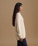 A woman in profile wearing a cream ribbed knit sweater and dark navy trousers, standing against a solid beige studio background.