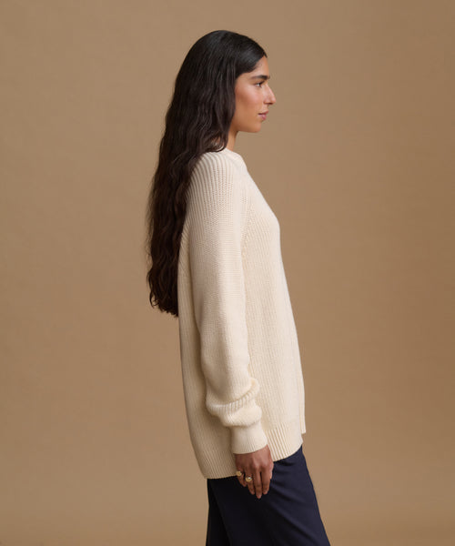 A woman in profile wearing a cream ribbed knit sweater and dark navy trousers, standing against a solid beige studio background.