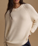 A close-up of a woman wearing a cream-colored ribbed knit sweater against a solid beige background.