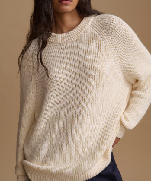 A close-up of a woman wearing a cream-colored ribbed knit sweater against a solid beige background.