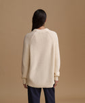 A woman viewed from the back wearing an oversized cream ribbed knit sweater and dark navy pants, standing against a solid warm tan studio background.