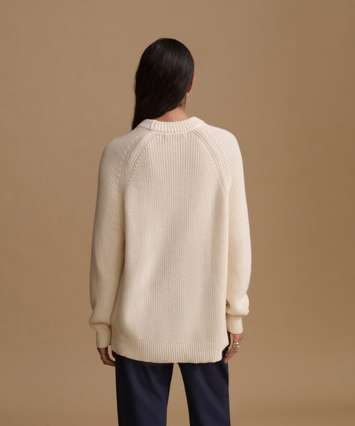 A woman viewed from the back wearing an oversized cream ribbed knit sweater and dark navy pants, standing against a solid warm tan studio background.