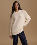 A woman modeling a cream ribbed knit sweater paired with dark navy pants, against a solid beige studio background.
