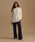 A woman stands against a warm beige studio background wearing a cream ribbed knit sweater, dark navy wide-leg trousers, and white pointed shoes.
