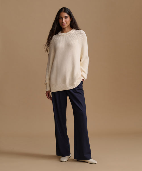 A woman stands against a warm beige studio background wearing a cream ribbed knit sweater, dark navy wide-leg trousers, and white pointed shoes.