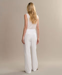 A woman with long blonde hair, wearing a sleeveless white top and the Paloma Pant, stands facing away from the camera against a beige background.