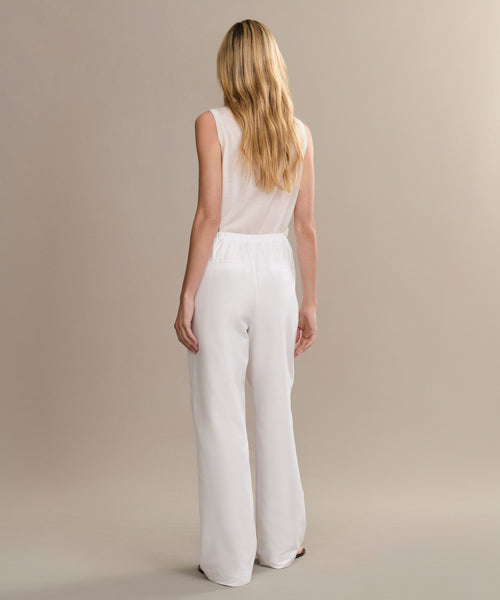 A woman with long blonde hair, wearing a sleeveless white top and the Paloma Pant, stands facing away from the camera against a beige background.