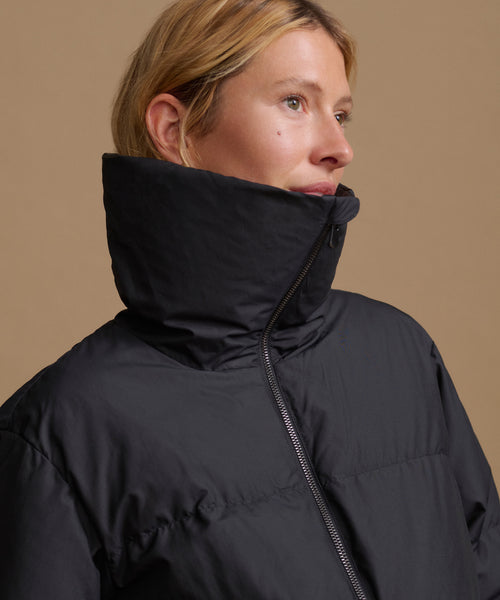 A close-up side profile of a woman wearing a black puffer jacket with a high collar, set against a solid warm beige background.