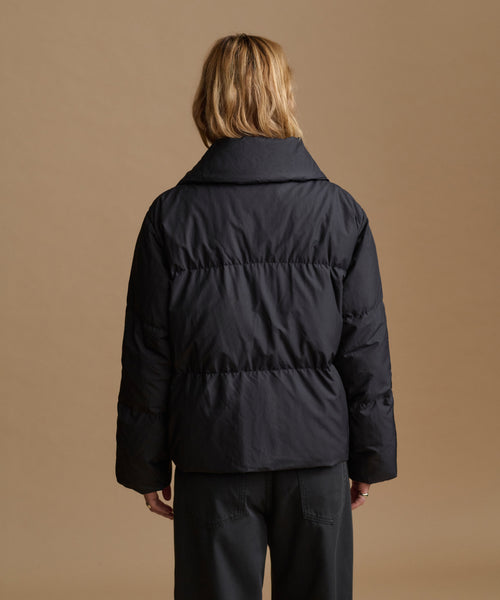 A model viewed from the back wearing a black puffer jacket and dark trousers, set against a solid warm tan studio background.